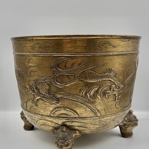 May include: A round brass incense burner featuring an embossed dragon motif wrapping around the exterior. The vessel is supported by three distinct feet shaped like stylized animal heads, with a weathered gold-toned finish and etched cloud patterns.