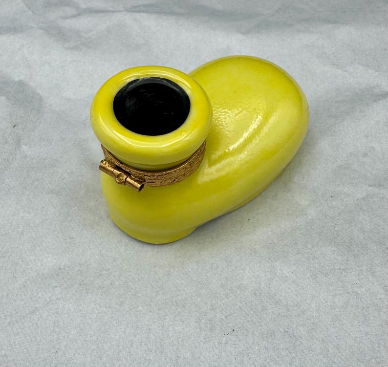 Pu&ograve; includere: Un posacenere in ceramica gialla con interno nero e una fascia metallica color oro. Il posacenere ha una forma curva unica ed &egrave; posto su uno sfondo neutro.