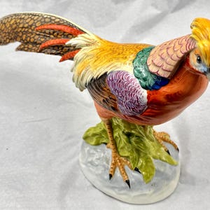 Op de afbeelding: Gedetailleerd keramiek beeldje van een fazant, met levendige kleuren. De vogel heeft een gele kuif, een oranje lichaam en een staart met bruine, rode en oranje patronen. Het staat op een voet met groene bladeren, een decoratief stuk.