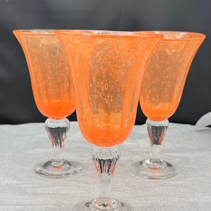 May include: Three orange-tinted wine glasses with clear, textured stems and bases. The glasses have a flared, tulip-shaped bowl. The glass has a bubbly texture. The glasses are arranged in a triangular formation.