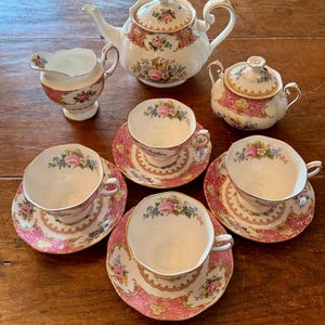 Puede incluir: Un juego de té vintage con tetera, cremera, azucarero y cinco tazas con platillos. El juego tiene una base blanca con detalles rosas y florales. Las tazas y platillos tienen aproximadamente 10 cm de diámetro.