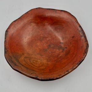May include: A shallow, round bowl with an irregular, wavy rim. The surface is a deep, mottled terracotta red with darker brown patches and subtle, swirling brushstroke textures visible across the interior basin of the vessel.