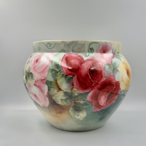 May include: A round porcelain bowl featuring a hand-painted floral design with large, vibrant red and soft pink roses set against a background of green leaves and muted foliage. The rim is finished with a thin, delicate gold-colored trim.