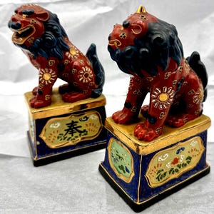 Puede incluir: Par de estatuas de leones guardianes de cerámica roja y negra. Las estatuas tienen detalles dorados y se asientan sobre pedestales azules con ribetes dorados y diseños florales. Los leones tienen una expresión feroz y están decorados con patrones florales.