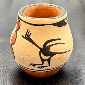 Puede incluir: Un pequeño jarrón de cerámica pintado a mano con un diseño de pájaro blanco y negro. El jarrón tiene un patrón geométrico rojo y blanco alrededor de la parte superior e inferior.