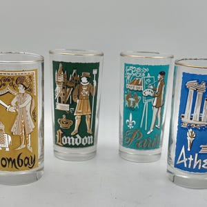 May include: Four clear glass tumblers with gold rims, each featuring a colored rectangular panel with a city name and illustration: Bombay in gold, London in green, Paris in teal, and Athens in blue, depicting cultural landmarks and figures.