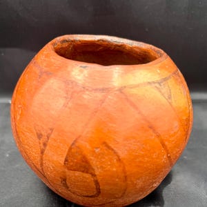 Puede incluir: Una pequeña olla de barro redonda y naranja con una abertura ancha y un patrón decorativo de líneas y curvas. La olla está hecha a mano y tiene un aspecto rústico y terroso.