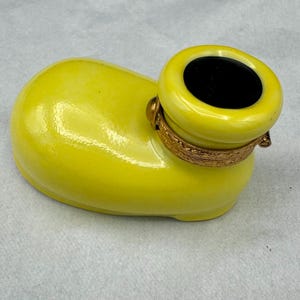 Pu&ograve; includere: Un contenitore in ceramica giallo brillante con un'apertura cilindrica e una fascia dorata decorativa. Il contenitore ha una finitura liscia e lucida e un interno nero. La forma generale ricorda una scarpa.