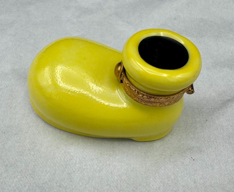 Pu&ograve; includere: Un contenitore in ceramica giallo brillante con un'apertura cilindrica e una fascia dorata decorativa. Il contenitore ha una finitura liscia e lucida e un interno nero. La forma generale ricorda una scarpa.