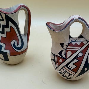 May include: Two small, hand-painted ceramic vases with Southwestern designs. One is a pitcher shape with a handle, and the other is a small, open-topped vase. Both feature red, black, and white geometric patterns.