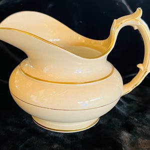 Puede incluir: Jarra de cerámica color crema con ribete dorado. La jarra tiene un cuerpo redondeado, un pico curvo y un asa decorativa. Bandas doradas rodean el cuerpo y el borde. Probablemente vintage o antigua.