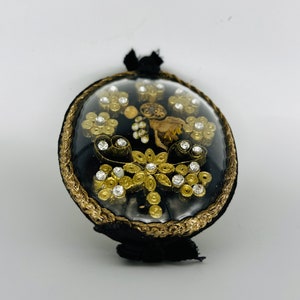 May include: A round, black velvet ornament with a clear, domed top. The ornament is decorated with gold-colored filigree, clear rhinestones, and small gold-colored floral accents. A gold-colored trim borders the clear dome. A black tassel is attached.
