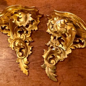 May include: Two ornate, gold-colored wall sconces with a classical design. The sconces feature intricate scrollwork and leaf patterns, creating a luxurious and antique aesthetic. The sconces are displayed on a wooden surface.