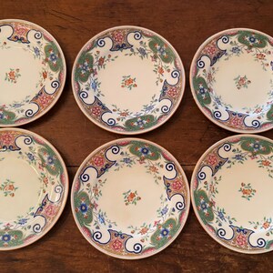 Puede incluir: Un juego de seis platos redondos de cerámica con un centro de color crema y un pequeño motivo floral. Los bordes anchos están decorados con un patrón intrincado de volutas azules, secciones verdes texturizadas y detalles florales rosas sobre fondo blanco.