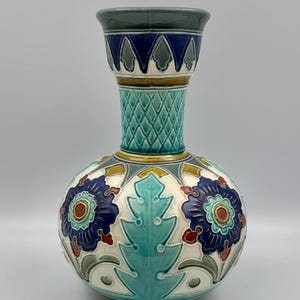 May include: A ceramic vase featuring a bulbous body and a flared neck. The surface is decorated with a relief pattern of dark blue flowers, teal leaves, and cream-colored accents. The neck has a cross-hatched texture and a dark blue rim.