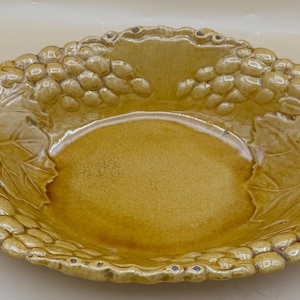 May include: Oval-shaped ceramic serving dish in a warm, golden-yellow hue. The rim is adorned with a grape cluster and leaf design. The dish has a smooth, glossy interior and a textured exterior, perfect for serving fruit or snacks.