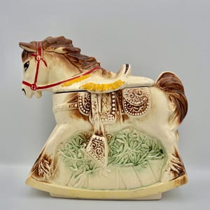 Pode incluir: Um pote de biscoitos de cavalo de baloiço de cerâmica em creme, castanho e vermelho. O cavalo tem uma sela e uma crina detalhadas, com um desenho de folhas verdes na base. O pote foi concebido para se assemelhar a um brinquedo vintage.