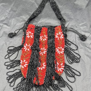 Può includere: Una borsetta di perline con manico e frange nere. La borsa presenta strisce rosse verticali con motivi floreali bianchi, separate da strisce di perline nere. Anche le frange e il manico sono neri. Un accessorio elegante.