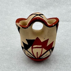 May include: A small, hand-painted ceramic vase. The vase is beige with red and black geometric designs. The rim and base are red, and it has a central hole and two smaller openings. The vase is likely of artisan origin.