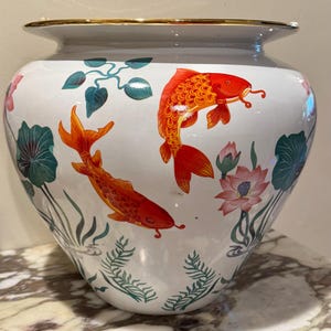 Könnte beinhalten: Weiße Keramikvase mit goldenem Rand, verziert mit orangefarbenen Kois und Illustrationen von Wasserpflanzen. Die Vase hat einen breiten Körper und eine leicht ausgestellte Oberseite. Das Design umfasst rosa Lotusblumen und grünes Laub.