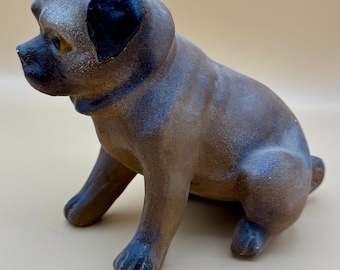 May include: A ceramic pug dog figurine in a seated position. The dog has a brown body with darker brown and black accents on the ears, nose, and paws. The figurine has a slightly glossy finish and is set against a neutral background.
