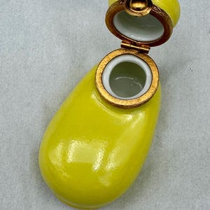 Pu&ograve; includere: Un portagioie in ceramica gialla con coperchio incernierato. La scatola ha una forma arrotondata con bordo e dettagli color oro. Il coperchio &egrave; aperto, rivelando un interno bianco. La scatola &egrave; su una superficie grigio chiaro.