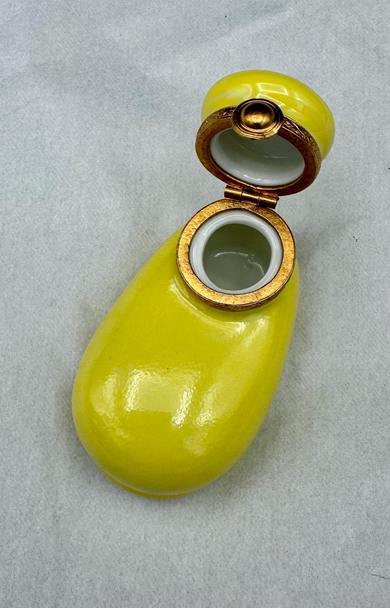 Pu&ograve; includere: Un portagioie in ceramica gialla con coperchio incernierato. La scatola ha una forma arrotondata con bordo e dettagli color oro. Il coperchio &egrave; aperto, rivelando un interno bianco. La scatola &egrave; su una superficie grigio chiaro.