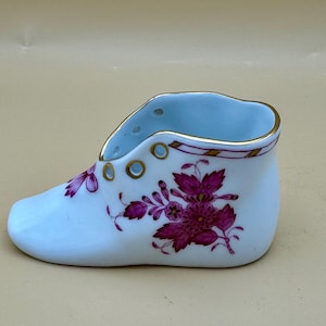 May include: A small, porcelain shoe-shaped decorative item. The shoe is primarily white with a light blue interior and floral designs in shades of pink and purple. Gold accents are present around the top rim and eyelets.