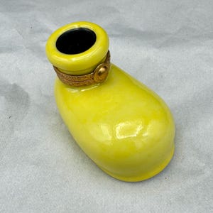 Pu&ograve; includere: Un contenitore decorativo in ceramica giallo brillante con interno nero. Il contenitore ha una forma arrotondata con una fascia metallica color oro attorno all'apertura. Il contenitore &egrave; su una superficie bianca.