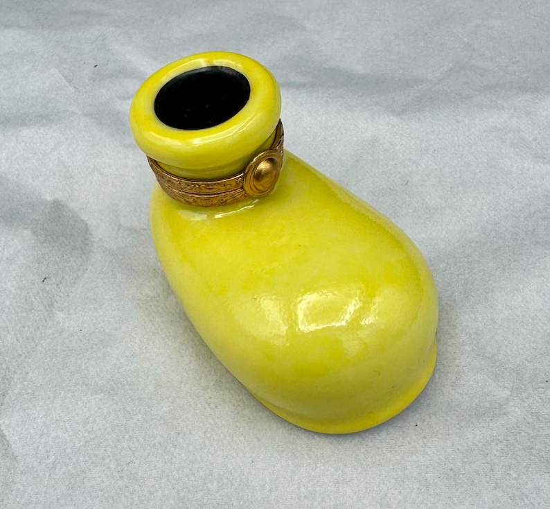 Pu&ograve; includere: Un contenitore decorativo in ceramica giallo brillante con interno nero. Il contenitore ha una forma arrotondata con una fascia metallica color oro attorno all'apertura. Il contenitore &egrave; su una superficie bianca.
