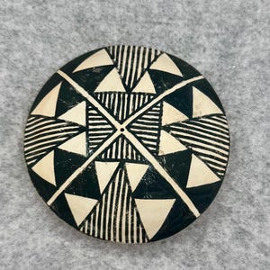 May include: A round, black and white decorative object with a geometric design. The design features triangles, lines, and a central point. The object is likely made of ceramic or wood.