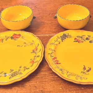 May include: A set of four yellow ceramic dishes. Two bowls with handles and two oval plates. Each piece is decorated with a floral and fruit design in red, green, and blue. The rims have a scalloped edge.