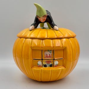May include: A ceramic pumpkin-shaped jar with a lid featuring a sculpted figure wearing a green hat and black clothing. The jar has a square window cutout on the front, revealing a painted figure inside with blonde hair and a green hair accessory.