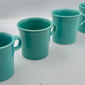 May include: Four matching turquoise ceramic mugs arranged in a diagonal line. Each mug features a smooth, glossy finish, a cylindrical body with a slightly flared rim, and a single curved handle on the side. The design includes subtle horizontal rings.