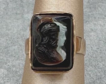 Op de afbeelding: Een gouden ring met een rechthoekige zwarte camee met twee gebeeldhouwde profielen. Eén figuur is donker, de andere licht, beide dragen hoeden. De ring wordt op een vinger getoond.