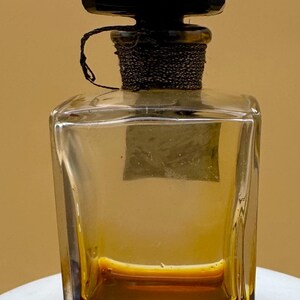 May include: A clear, rectangular glass perfume bottle with a dark brown liquid inside. The bottle has a black, geometric stopper and a decorative, dark brown chain around the neck. The bottle sits on a white surface.