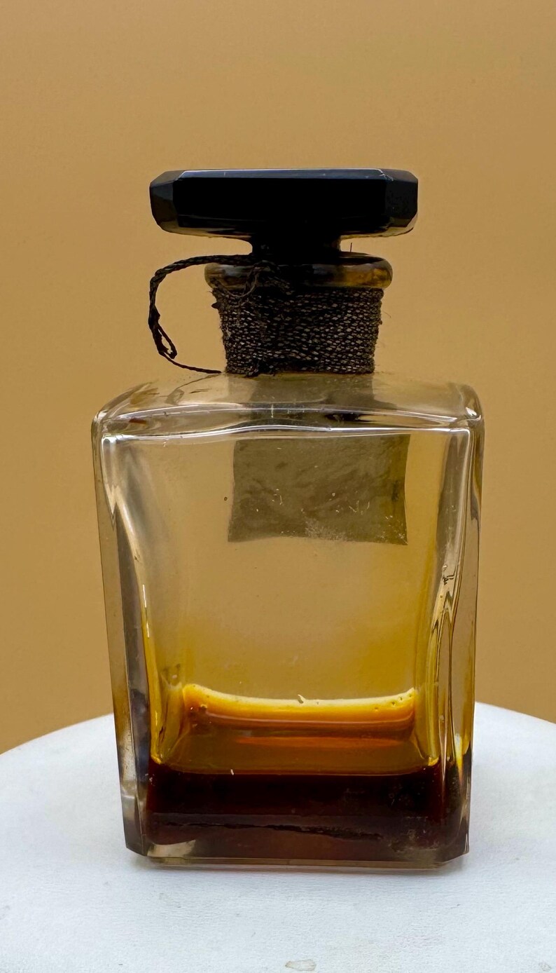 May include: A clear, rectangular glass perfume bottle with a dark brown liquid inside. The bottle has a black, geometric stopper and a decorative, dark brown chain around the neck. The bottle sits on a white surface.