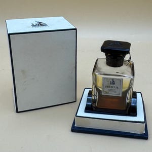May include: A perfume bottle with a black cap and a label that reads "Pre-Texte Lanvin Paris" sits on a white and black stand. The bottle is next to a white box with a black trim and a logo on the top.