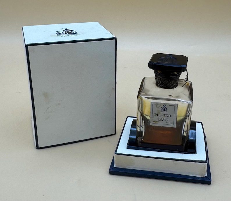 May include: A perfume bottle with a black cap and a label that reads "Pre-Texte Lanvin Paris" sits on a white and black stand. The bottle is next to a white box with a black trim and a logo on the top.