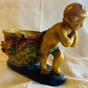 Op de afbeelding: Een gouden cherubfiguur dat een mand met druiven en bladeren draagt. De mand is versierd met een roze lint en groene en bruine bladeren. De cherub staat op een blauwe en zwarte basis.
