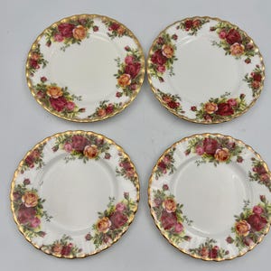 May include: A set of four white ceramic plates with scalloped edges and gold trim. Each plate features a floral pattern of red and yellow roses with green leaves arranged along the rim. The plates have a smooth, glossy finish and circular shape.