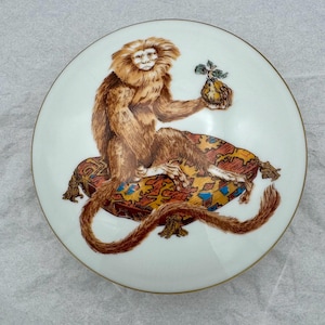 May include: A round, white decorative plate with a detailed illustration of a monkey seated on a patterned cushion. The monkey is brown with a light-colored face and is holding a fruit. The plate has a gold rim.