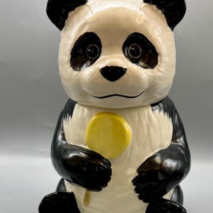 Pode incluir: Um pote de biscoitos de cerâmica em forma de panda com pelo preto e branco. O panda está segurando uma pirulito amarelo na pata direita. O pote tem uma tampa e é uma adição divertida e caprichosa a qualquer cozinha.