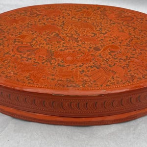 May include: A round, red lacquered box with a detailed, embossed design on the lid. The lid features a pattern of various figures and shapes in a darker shade of red. The sides of the box have a repeating, curved design.