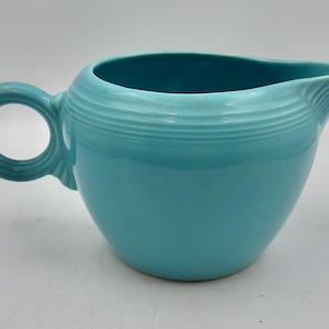 May include: A turquoise ceramic creamer jug featuring a rounded body, a circular handle, and a prominent pouring spout. The exterior surface is decorated with a series of horizontal concentric rings encircling the upper portion of the vessel.