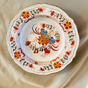 May include: White ceramic plate with a scalloped edge and floral design. The plate features a central floral arrangement in red, yellow, and blue, surrounded by a border of smaller flowers and green leaves. The rim has a gold trim.
