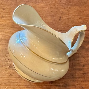 Puede incluir: Jarra de cerámica color crema con pico y asa curvados. La jarra presenta adornos dorados y un diseño en espiral. El asa tiene un diseño en forma de rama. La jarra está sobre una superficie de madera. Medidas en cm.