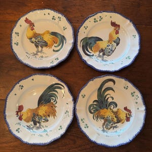 Peut inclure: Quatre assiettes en céramique blanche avec un bord festonné et une bordure bleue. Chaque assiette présente une illustration colorée de coq en jaune, orange, bleu et vert. Les assiettes ont un style rustique et champêtre.