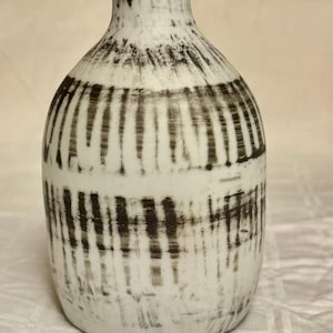 May include: A white ceramic vase with a rounded body and a narrow neck. The vase is decorated with vertical and horizontal black brushstrokes, creating a textured, abstract pattern. The vase is approximately 8 inches tall.