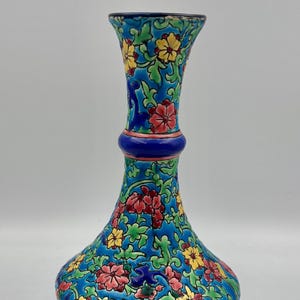 May include: A decorative vase featuring a vibrant blue background with an intricate pattern of red and yellow flowers intertwined with green foliage. The vase has a narrow neck, a bulbous body, and a solid dark blue base and rim.